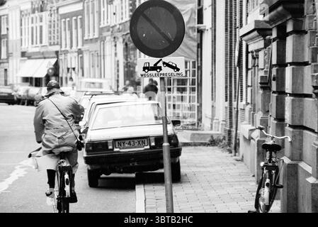 Parkplatzbelästigung Jant@@ [Jansstraat], Whizgle News, Dutch Desk, Niederlande, 1950 - 2000 am 03-06-1991. Diese Themen werden in der Abbildung angezeigt. Die Szene zeigt eine geschäftige urbane Straße, die in Schwarzweiß gehalten wird, was ihr eine zeitlose Qualität verleiht. Im Vordergrund fährt ein Radfahrer am Straßenrand entlang und trägt eine helle Regenjacke, die sich von den dunkleren Farben der Umgebung abhebt. Der Radfahrer scheint fokussiert zu sein und deutet auf einen Sinn für einen Zweck auf seiner Reise hin. Neben der Straße ist ein geparktes Auto zu sehen, dessen schlanke Form teilweise vom Radfahrer verdeckt wird. Das Fahrzeug Stockfoto