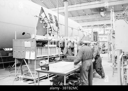 Produktionshalle Fokker F28 Fokker, Whizgle News, Dutch Desk, Niederlande, 1950 - 2000 am 01-03-1979. Das Bild enthält diese Themen. Die Szene zeigt eine industrielle Umgebung, wahrscheinlich innerhalb einer Montage- oder Wartungseinrichtung für Flugzeuge. Im Vordergrund sind mehrere Personen mit Arbeitsaufgaben beschäftigt, alle in dunklen, zweckmäßigen Overalls gekleidet, was auf Funktionalität und Sicherheit hinweist. Die Arbeiter konzentrieren sich auf verschiedene Aktivitäten rund um einen großen, teilweise gebauten Flugzeugrumpf, der die linke Seite der Szene dominiert. Seine metallische Oberfläche reflektiert das Umgebungslicht Stockfoto