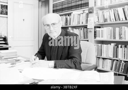 Bischof Bomers of Haarlem Roman Catholic Church Haarlem the Netherlands, Whizgle News, Dutch Desk, Niederlande, 1950 - 2000 am 03-09-1984. Diese Themen werden in der Abbildung angezeigt. Die Szene zeigt einen schwach beleuchteten Raum mit Büchern und Papieren, der eine akademische Atmosphäre vermittelt. Im Vordergrund sitzt ein Mann in einem formellen schwarzen Anzug mit einem knusprigen weißen Hemd an einem Schreibtisch, was ein Gefühl von Professionalität und Konzentration schafft. Er hat einen nachdenklichen Ausdruck, der Konzentration und Kontemplation suggeriert. Um ihn herum befinden sich Stapel von Büchern und verstreuten Dokumenten, die auf ein geschäftiges und möglicherweise sogar ein geschäftiges Verhalten hinweisen Stockfoto