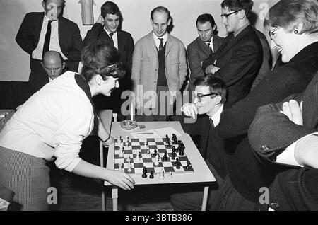 Fünf Damen vom Hoogoven Chess Tournament - Leser IJmuider Courant Chess, Whizgle News, Dutch Desk, Niederlande, 1950 - 2000 am 22.01.1965. Das Bild enthält diese Themen. In einem belebten, von Kameradschaft und Fokus geprägten Innenbereich versammelte sich eine Gruppe von Menschen um einen Schachtisch. Die Szene ist von einer Mischung aus Neugier und Spannung erfüllt, während zwei Individuen tief in ein Schachspiel verwickelt sind. Eine junge Frau in einem hellen Oberteil und einer gemusterten Hose beugt sich leicht nach vorne am Tisch. Ihr Ausdruck zeigt Konzentration und Entschlossenheit als Stockfoto
