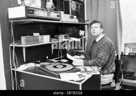 Lokaler Radio DJ, Whizgle News, Dutch Desk, Niederlande, 1950 - 2000 am 02-1990. Dies sind die Themen im Bild. In einem gemütlichen Aufnahmestudio sitzt ein Mann an einem überdachten Schreibtisch, der mit verschiedenen Musikgeräten und Erinnerungsstücken geschmückt ist. Der Hintergrund ist mit einer reichhaltigen Textur aus Vorhangstoff versehen, die der Szene ein warmes, intimes Gefühl verleiht. Der Schreibtisch, der hauptsächlich aus dunklem Holz gefertigt ist, zeigt eine Reihe von Audiogeräten: Einen Plattenspieler, der in der Mitte deutlich positioniert ist und auf dem eine Schallplatte sanft aufliegt. Um den Drehtisch herum befinden sich Stapel von Fotos und kleine Papierstücke, hintin Stockfoto