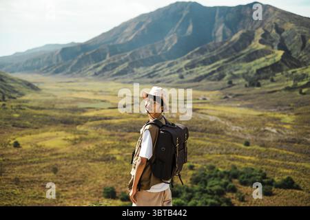 Ein südostasiatischer Mann mit Brille, Eimerhut und braunem Rucksack lächelt, während er zurück in die Kamera blickt. Er steht in einem riesigen grasbewachsenen Tal mit Stockfoto