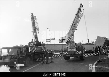 Schwere Last auf einem Anhänger mit Auflieger..., Whizgle News, Dutch Desk, Niederlande, 1950 - 2000. Dies sind die Themen im Bild. Die Szene zeigt eine geschäftige Baustelle, die sich um zwei große Kräne dreht, die an einer Fahrbahn platziert sind. Die Kräne, hoch- und Industriekrane, sind hoch angewinkelt, ihre Arme ausgestreckt, während sie eine massive Betonplatte in die Luft heben. Die Karosserie der Kräne zeigt eine Mischung aus metallischem Grau und hellgelbem, was ihr robustes Design und ihre Funktionalität bei klarem Himmel unterstreicht. Vor Ort ist eine Gruppe von Arbeitnehmern an Koordinaten beteiligt Stockfoto