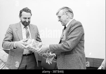 Abschied Cor Hoogland IJmuiden (Pilotendienst) Pilotenpiloten IJmuiden Niederlande, Whizgle News, Dutch Desk, Niederlande, 1950 - 2000 am 06-04-1982. Das Bild enthält diese Themen. Die Szene zeigt zwei Männer, die in einem Moment des Geschenks verwickelt sind, beide in formeller Kleidung. Ein Mann mit einem Bart und einem freundlichen Ausdruck überreicht ein hell gewickeltes Geschenk mit einem komplizierten Muster, das in leuchtenden Farben und geometrischen Formen verziert ist. Der andere Mann trägt eine Brille und einen Anzug mit einem dezenten karierten Muster und zeigt einen Blick der Wertschätzung und Neugier, während er die Prägung empfängt Stockfoto