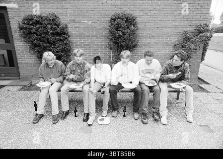 Barbecue Technical School IJm. IJmuiden the Netherlands, Whizgle News, Dutch Desk, Niederlande, 1950 - 2000 am 17-06-1993. Dies sind die Themen im Bild. Eine Gruppe von sechs jungen Individuen sitzt auf einer einfachen Bank, die in einer Reihe ausgerichtet ist. Sie scheinen ein gemeinsames Essen zu genießen. Die Kulisse bietet eine strukturierte rote Backsteinwand, die der Szene Wärme verleiht. Jede Person ist lässig gekleidet, mit verschiedenen Mustern und Farben in ihren Hemden, was zu einer entspannten und ungezwungenen Atmosphäre beiträgt. Ihre Frisuren sind unterschiedlich und spiegeln Jugendliche Individualität und vielleicht jüngste tr wider Stockfoto