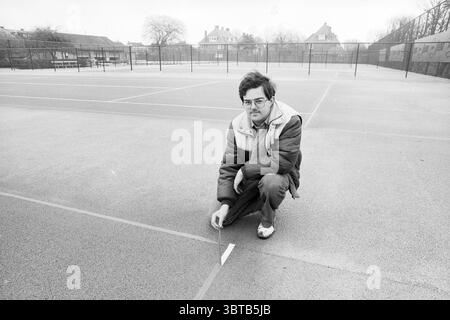 Portrait man at Tennis Court., Whizgle News, Dutch Desk, Niederlande, 1950 - 2000. Die Abbildung zeigt diese Themen. In einer kargen Umgebung im Freien kniet ein Mann auf einer grauen, strukturierten Oberfläche, wahrscheinlich auf einem Tennisplatz. Er trägt eine dicke, dunkle Jacke, die sich über hellerer Kleidung befindet und ein Gefühl von lässiger Zweckmäßigkeit ausstrahlt. Seine Brille und sein leicht ungepflegtes Haar deuten auf ein nachdenkliches Verhalten hin, während er sich intensiv auf eine Linie konzentriert, die auf dem Platz markiert ist. Die Kulisse zeigt eine Reihe von einfachen Gebäuden, die mit einer Mischung aus flachen Dächern und rechteckigen Formen eher zweckmäßig wirken. Diese Strukturen sind Stockfoto