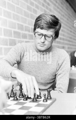 Head of Chess Players Blast Furnace Chess Tournament, Whizgle News, Dutch Desk, Niederlande, 1950 - 2000 am 11-01-1972. Das Bild enthält diese Themen. Ein junger Mann sitzt an einem kleinen Tisch und konzentriert sich intensiv auf ein Schachspiel. Er hat ordentlich dunkles Haar und trägt eine große, runde Brille, die seine Konzentration unterstreicht. Sein Pullover ist in sanftem Grau gehalten, das sich mit den gedämpften Tönen des Setups harmoniert. Vor ihm befindet sich ein klassisches Schachbrett, dessen Stücke in ihren Ausgangspositionen angeordnet sind und sich scharf gegen die helle Oberfläche des Bretts abheben. Die Schachfiguren sind aus einem gefertigt Stockfoto