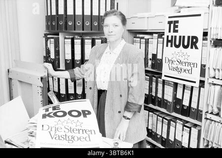 Mrs. Stolpe erste Immobilienmaklerin Makelaars, Whizgle News, Dutch Desk, Niederlande, 1950 - 2000 am 12.09.1989. Diese Themen werden in der Abbildung angezeigt. Die Szene zeigt eine Frau, die in einer Büroumgebung steht, die durch eine geschäftige Atmosphäre gekennzeichnet ist. Sie hat einen Ausdruck, der Entschlossenheit oder Konzentration vermittelt. Ihre Kleidung ist mit einem strukturierten Blazer über einer hellen Bluse versehen und sorgt für einen professionellen Auftritt. Um sie herum bilden mehrere große Aktenschränke eine Kulisse, gefüllt mit ordentlich organisierten Akten, die eine Fülle von Dokumentationen und möglicherweise ein großes Volumen suggerieren Stockfoto