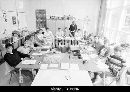 World Record class de Vries Mavo Record Improves and Improvement, Whizgle News, Dutch Desk, Niederlande, 1950 - 2000 am 26-04-1983. Das Bild enthält diese Themen. In einem großen Klassenzimmer sitzt eine Gruppe von rund 20 Schülern an Tischen, die in einer langen U-Form angeordnet sind. Die Farbpalette ist überwiegend gedämpft, mit Grautönen und einigen helleren Tönen, die auf eine Vintage- oder monochrome Atmosphäre hinweisen. Durch große Fenster strömt natürliches Licht ein, das weiche Schatten wirft und eine warme, aber fleißige Stimmung schafft. Die Schüler, eine vielfältige Mischung aus Geschlechtern und Frisuren, scheinen sich mit va beschäftigt zu sein Stockfoto