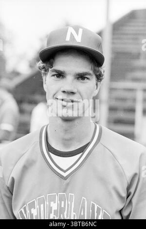 Spieler niederländisches Baseballteam Baseball, Whizgle News, Dutch Desk, Niederlande, 1950 - 2000 am 10-07-1984. Das Bild enthält diese Themen. In dieser Szene steht ein junger Mann zuversichtlich, trägt eine Baseballmütze, die mit einem kühnen Buchstaben verziert ist. Sein Haar ist hell und leicht gewellt, umrahmt sein Gesicht. Er hat einen freundlichen Ausdruck, mit einem leichten Lächeln, das sein offenes Auftreten unterstreicht. Er trägt ein Baseballtrikot mit lebhaften Farben, vor allem in Orange- und Weißtönen, die einen schönen Kontrast zum dezenten Hintergrund bilden. Das Trikot hat ein sportliches Design, das mit einer Naht hervorgehoben wird Stockfoto