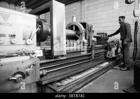 Mr. Leek und Dol Ned. Metal Foundries, Whizgle News, Dutch Desk, Niederlande, 1950 - 2000 am 26-12-1995. Das Bild enthält diese Themen. Die Szene fängt eine Werkstatt mit industrieller Atmosphäre ein. Im Vordergrund steht eine große Drehmaschine aus robustem Metall mit leicht abgenutzter Oberfläche, die auf eine umfangreiche Nutzung hinweist. Die Maschine wird von einem zylindrischen Werkstück dominiert, das sich gerade in Form befindet, mit verschiedenen Zifferblättern und Hebeln an der Seite, die auf seine technische Funktion hinweisen. Zur Seite steht ein Mann aufmerksam an der Drehmaschine. Er Stockfoto