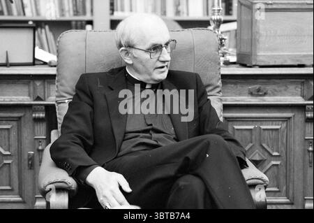 Bischof Bomers of Haarlem Roman Catholic Church Haarlem the Netherlands, Whizgle News, Dutch Desk, Niederlande, 1950 - 2000 am 03-09-1984. Diese Themen sind in der Abbildung dargestellt. Die Szene zeigt einen Mann mittleren Alters, der bequem in einem Sessel sitzt und formell in einem schwarzen Anzug mit klerikalem Kragen gekleidet ist, der einen Hauch von Autorität und Ruhe ausstrahlt. Er trägt eine rechteckige Brille, die zu seinem nachdenklichen Verhalten beiträgt. Seine Körpersprache ist entspannt, ein Bein wird über das andere gekreuzt, und er lehnt sich leicht nach vorne, was auf ein Gespräch hinweist. Das Ambiente ist ein warm eingerichtetes Zimmer, charakteristisch Stockfoto