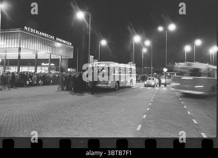 Ersatz-NZH-Bus-Transport für den Zug., Whizgle News, Dutch Desk, Niederlande, 1950 - 2000. Das Bild enthält diese Themen. Die Szene fängt nachts eine geschäftige urbane Umgebung ein, die von Straßenlaternen beleuchtet wird, die ein sanftes Leuchten über die Gegend werfen. Im Vordergrund steht ein Oldtimer-Bus, dessen Metallfläche das gelbliche Licht von oben reflektiert. Seitlich ist ein kleines Auto in Bewegung, dessen Scheinwerfer die Straße beim Vorbeifahren beleuchten. Die Umgebung ist mit einer Mischung aus architektonischen Elementen gestaltet – ein modernes Gebäude mit großen Glasfenstern ist hervorzuheben, während sich die Menschenmenge versammelt Stockfoto