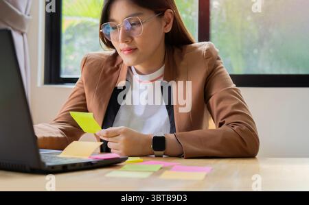Asiatische Geschäftsfrau mit Laptop und Blick auf eine Post-IT-Notiz zur Hand und klug im Büro. Porträt-Konzept. Stockfoto