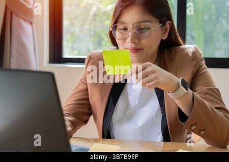 Lächelnde asiatische Geschäftsfrau, die sich eine Post-IT-Notiz auf ihrer Hand und ihrem Verstand ansieht. Arbeitende Frau, Unternehmensführungskonzept. Stockfoto