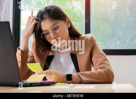 Nachdenkliche asiatische Frau mit langen Haaren in Brille, die an einem Schreibtisch arbeitet, umgeben von bunten Post-it-Notizen. Brainstorming-Konzept. Stockfoto