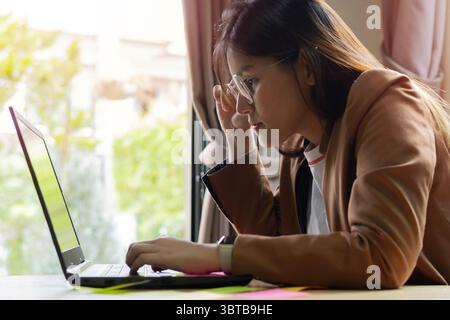 Junge, schöne asiatische Frau mit Brille, die im Büro am Computer arbeitet. Geschäftskonzept. Seitenansicht. Stockfoto