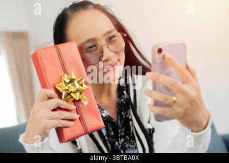 Asiatische Frau mittleren Alters mit Smartphone Nehmen Sie ein Selfie mit Weihnachtsgeschenk, während Sie zu Hause auf dem Sofa sitzen. Stockfoto