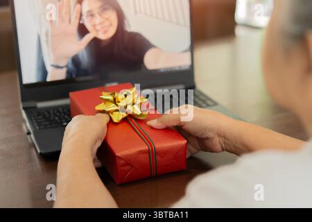 Eine ältere Frau, die Video-Chat hat, nachdem sie ein Geschenk von ihrer Familie erhalten und ein weihnachtsgeschenk ausgepackt hat. Rückansicht, selektiver Fokus. Stockfoto