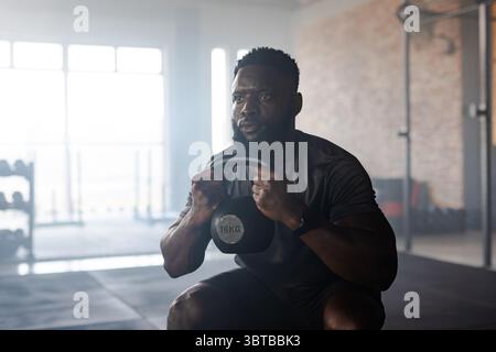 Afroamerikaner, der 16 kg Kettlebell hält, hockt auf Gummiboden im Fitnessstudio mit Hantelständer Stockfoto