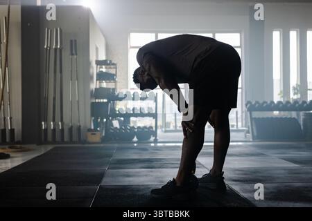 Afroamerikanischer Mann mit Sportbekleidung, der Atem schnappt und sich auf die Knie im Fitnessstudio an Gewichtsträgern lehnt Stockfoto