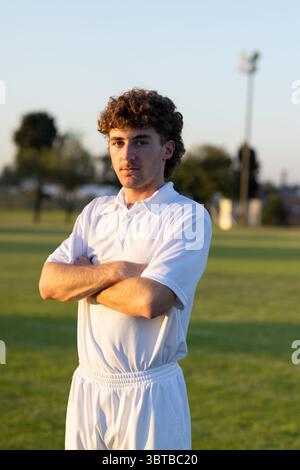 Erwachsener männlicher Athlet, der mit weißem Polo und Hose unter dem Flutlichtturm auf dem Cricketfeld steht Stockfoto