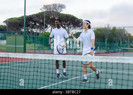 Verschiedene männliche Freunde in Sportbekleidung feiern den Sieg auf dem grünen Tennisplatz per Netz und halten Schläger Stockfoto