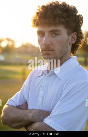 Mann, der bei Sonnenuntergang auf einem grasbewachsenen Sportplatz steht, ein Hemd mit Kragen trägt und einen Ohrring zeigt Stockfoto