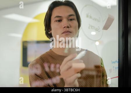Asiatischer Mann, der kreisförmige Diagramme auf Klarglas-Whiteboard im Büro mit Marker und Haftnotiz zeichnet Stockfoto
