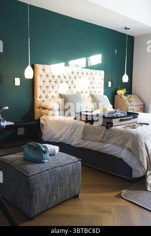 Das getuftete beige Bett ist mit offenem Koffer und gemusterten Kissen im blauen Hauptschlafzimmer ausgestattet Stockfoto