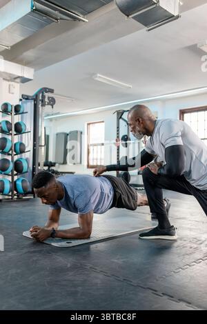 Afroamerikanischer Trainer und Kunde, der Unterarmplanke auf Matte im Fitnessstudio mit Hockgestell vorführt Stockfoto