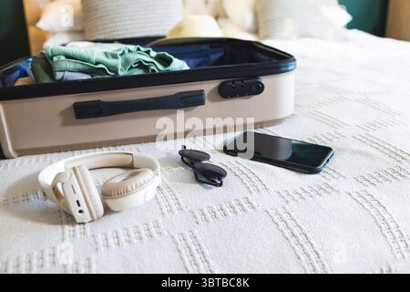 Offener Koffer, der auf dem Hotelbett liegt, mit gefalteter Kleidung, Strohhut, Kopfhörern, Sonnenbrillen, Smartphone Stockfoto
