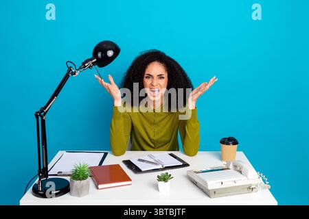 Frustrierte junge Frau, die am Schreibtisch mit Requisiten auf blauem Hintergrund arbeitet und Emotionen ausdrückt Stockfoto