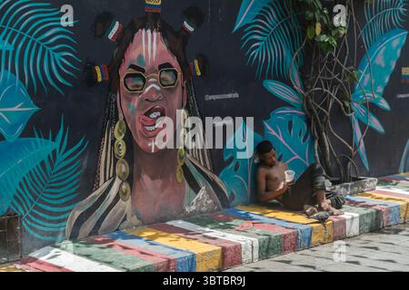Obdachloser Mann, der sich an eine Wand lehnt, mit einem Wandgemälde eines Mädchens mit einer Sonnenbrille, die ihre Lippen leckt. Getsemani, Cartagena, Departement Bolívar, Kolumbien Stockfoto