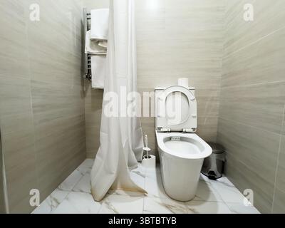 Eine weiße Toilette ist in einem Badezimmer mit Duschvorhang. Es gibt zwei Handtücher, die auf einem Regal hängen Stockfoto