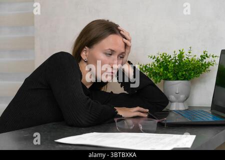 Die erschöpfte Freiberuflerin berührt ihren Kopf vor Schmerzen, während sie zu Hause an einem Laptop arbeitet und mit Burnout und projektbezogenem Stress zu kämpfen hat Stockfoto