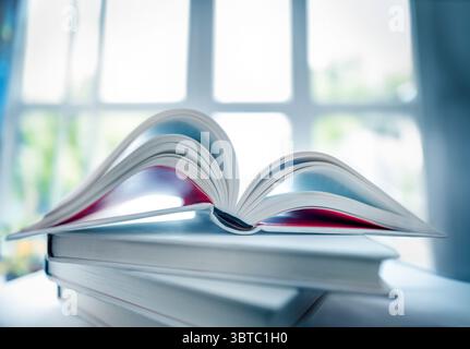 Flacher Fokus auf einen Stapel von Büchern und weit geöffnetes Buch darauf in einer angenehmen Atmosphäre zu Hause mit einem hellen Fenster und unfokussiertem Hintergrund Stockfoto