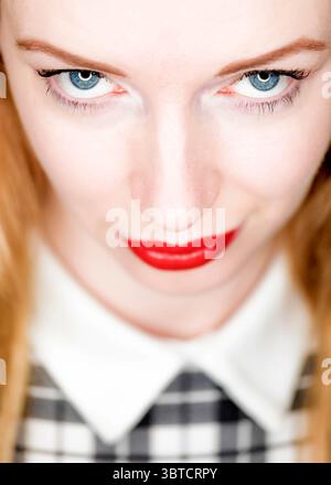 Eine Nahaufnahme des Gesichts einer Frau, fokussiert auf ihre Augen und Lippen. Sie hat blaue Augen, roten Lippenstift und trägt ein weißes Hemd mit Kragen und ein schwarzes Stockfoto