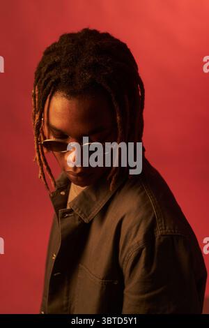 Schwarzer junger erwachsener Mann mit Sonnenbrille, der mit ernstem Ausdruck vor rotem Hintergrund nach unten blickt, Dreadlocks sichtbar, Kopf leicht von der Kamera abgewandt Stockfoto
