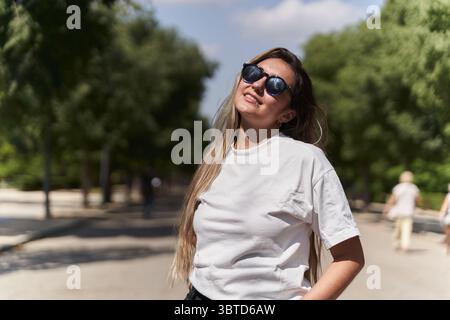 Glückliche junge Frau, die Sonnenbrille trägt und Sonnenlicht in einem Park genießt und unbeschwerte und positive Emotionen ausdrückt Stockfoto