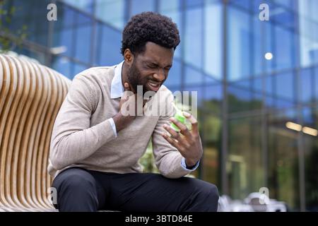Ein afroamerikanischer Mann leidet unter Halsschmerzen und benutzt draußen ein Kehlspray, in der Nähe eines modernen Glasgebäudes. Stockfoto