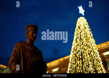 Eine Bronzestatue steht in Pamplona, Navarra, España, mit einem wunderschön beleuchteten Weihnachtsbaum, der hell vor dem tiefen Abendhimmel leuchtet. Das fest Stockfoto