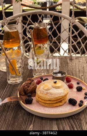Ein gemütlicher Brunch im Freien mit gestapelten Pfannkuchen, die mit Puderzucker, frischen Beeren und warmen Tees auf einem rustikalen Tisch bestreut sind Stockfoto