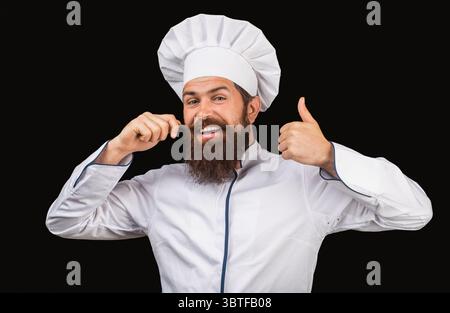 Glücklicher Koch, junger Mann mit Bart, der Daumen nach oben zeigt, lächelnd. Kochmütze. Bärtige Köche, Köche oder Bäcker positive Geste mit der Hand, Daumen hoch Stockfoto