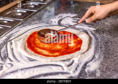 Prozess der Zubereitung von Pizza. Der Koch gießt Tomatensauce über den gerollten Teig. Chef Hand verteilt pasteurisierte Tomatenpaste auf eine Pizzabasis. Nahaufnahme Stockfoto