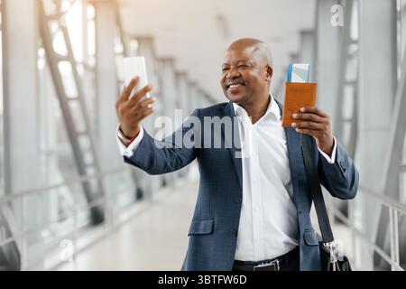 Glücklicher Geschäftsmann, der Selfie mit Smartphone am Flughafen macht, während er seinen Pass hält Stockfoto