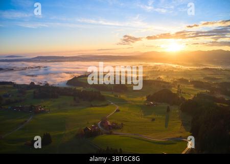 Aus der Vogelperspektive auf den Sonnenaufgang über dem Horizont, die Landschaft mit goldenem Licht, während Nebel tief über sanften grünen Feldern hängt, Bernbeuren, Bayern, Deutschland. Stockfoto