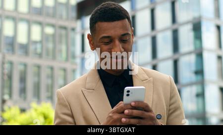 Glücklicher afroamerikanischer ethnischer Geschäftsmann in der Stadt im Freien Bürogebäude Arbeitgeber Geschäftsmann Mann männlicher Unternehmer, der scrollendes Handy durchsucht Stockfoto
