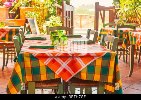 Restauranttische im Freien mit farbenfrohen karierten Tischdecken und grünen Gläsern für Gäste unter warmer mediterraner Sonne, die eine gemütliche und farbige Atmosphäre schaffen Stockfoto