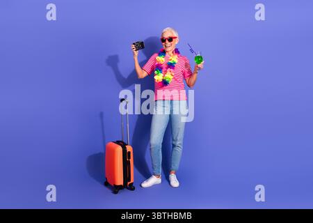Seniorin genießt Urlaubsstimmung in farbenfroher Kleidung, hält eine Kamera und trinkt vor einem hellen violetten Hintergrund Stockfoto